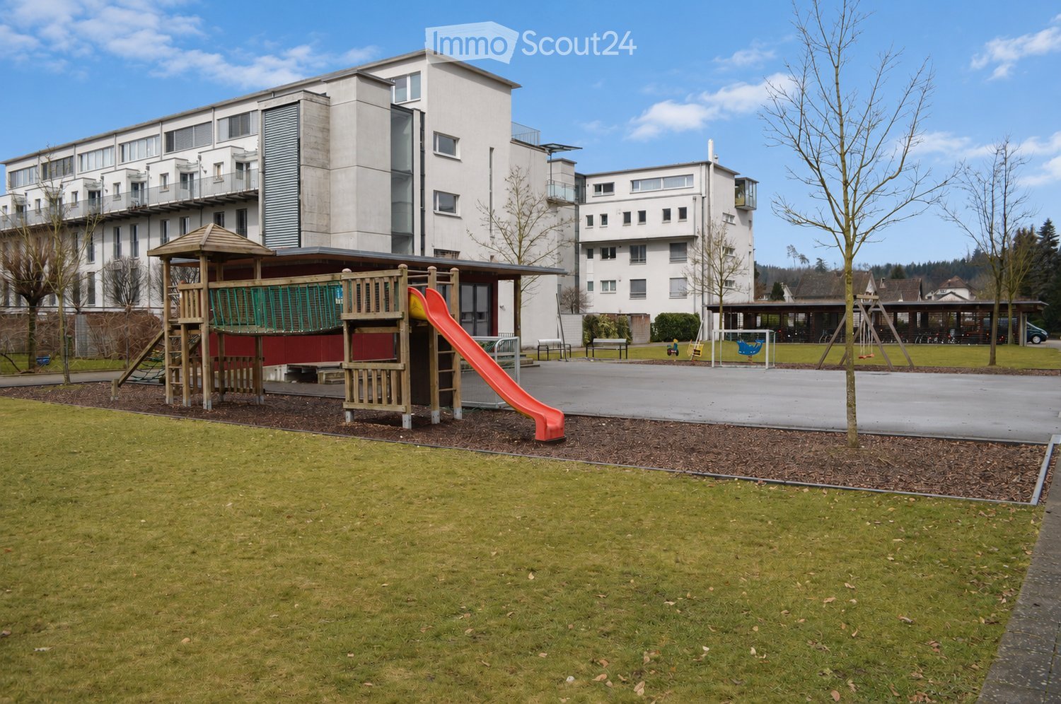 A modern residential building with balconies and a playground area featuring a slide, climbing structure, and benches.