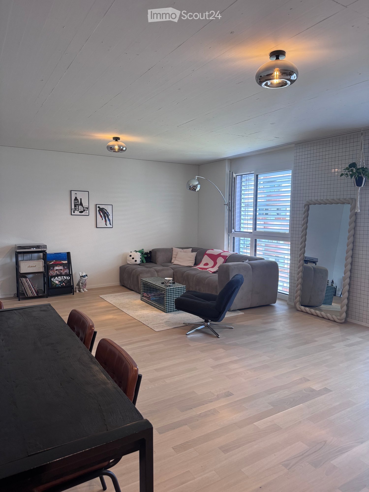 Modern living room with wooden flooring, large windows, a grey sectional sofa, a glass coffee table, a blue armchair, a dark wooden dining table, and a shelf with books and a small decorative item.