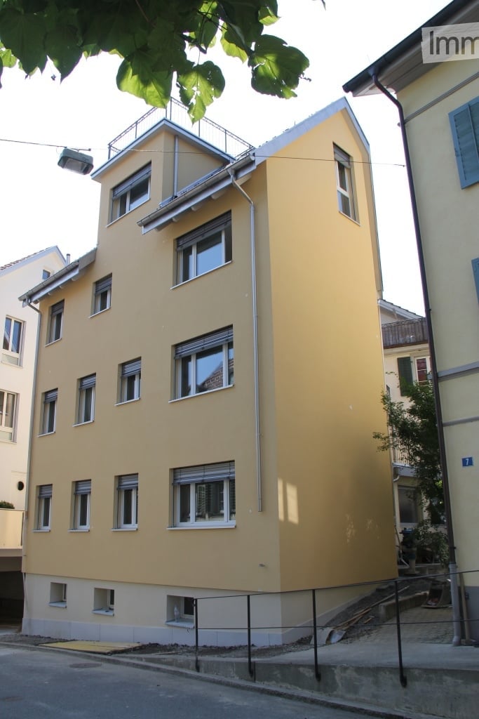 Four-story yellow building with multiple windows and a raised ground floor, surrounded by neighboring structures and greenery.