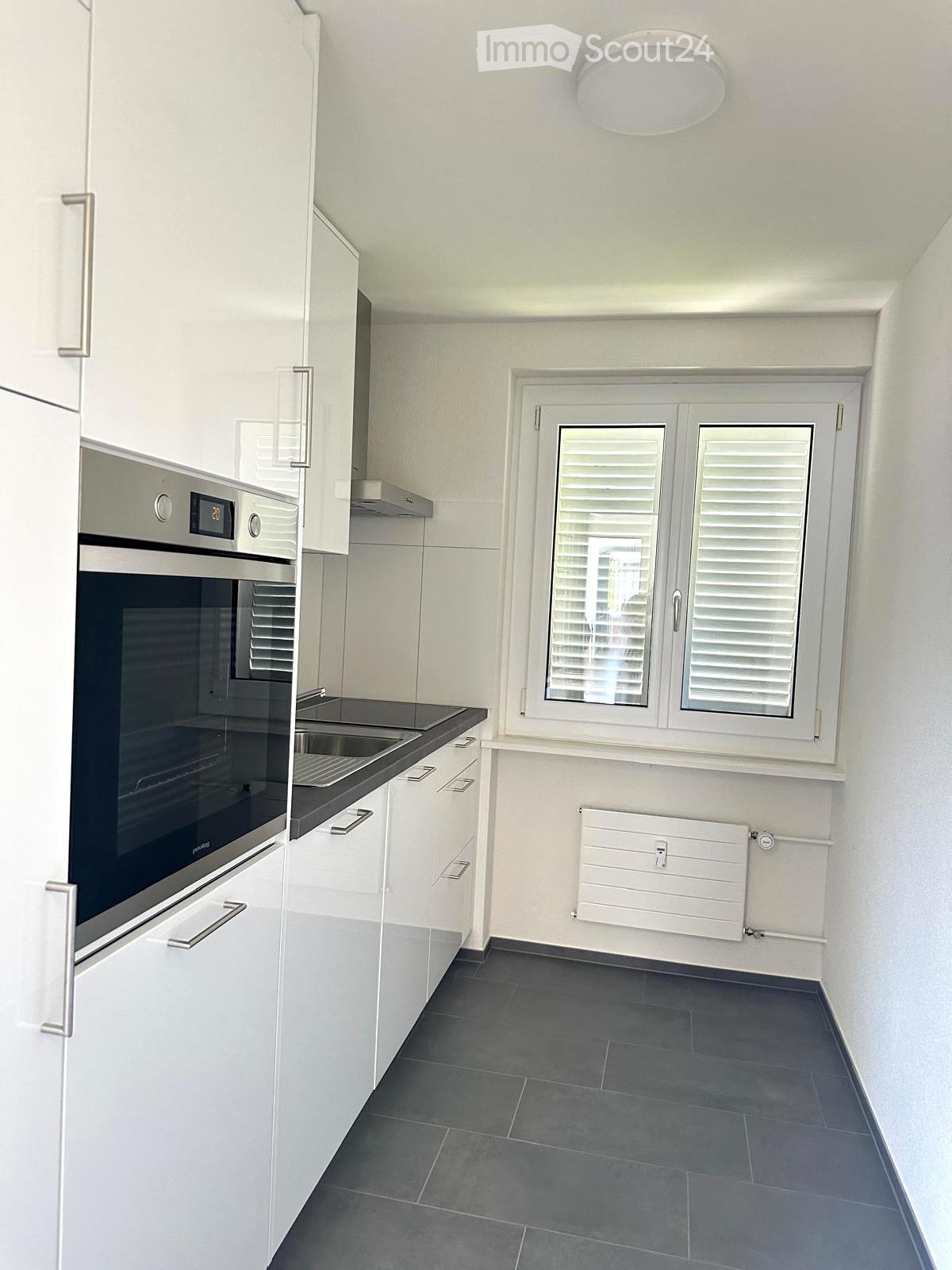 Modern kitchen with white cabinets, stainless steel oven, sink, and a window with shutters. Grey tiled floor and white walls.