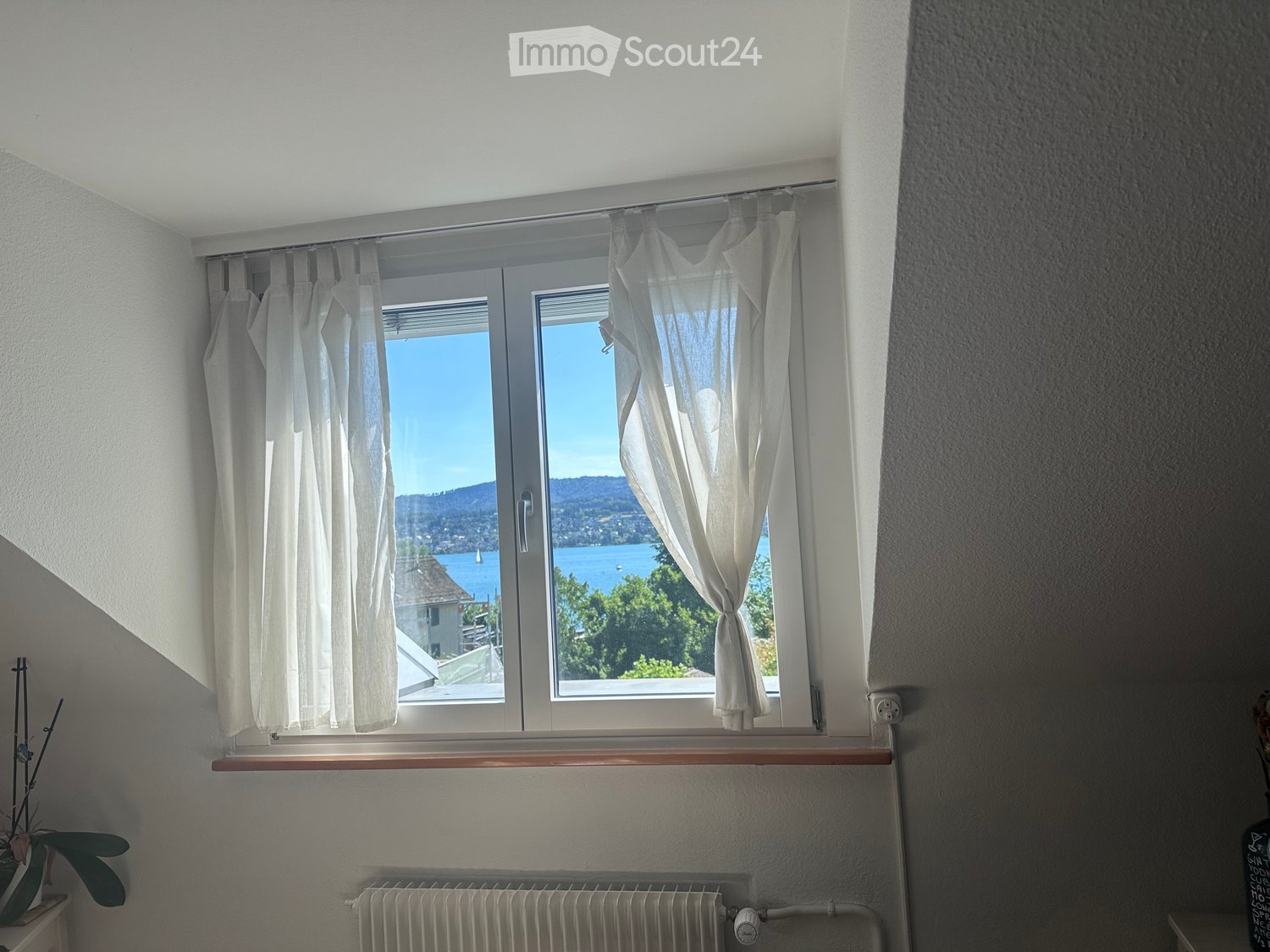 An unfurnished room with large windows offering a scenic view of a lake and surrounding landscape. The room features white walls, a radiator, and a wooden window sill.