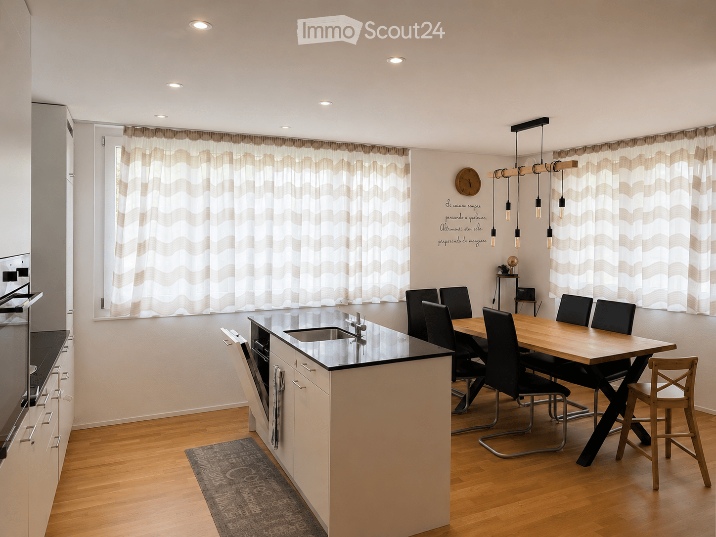 Modern kitchen and dining area with a central island, sink, and cabinets. A wooden dining table with black chairs is positioned near large windows with beige curtains. A small rug is placed on the floor near the island.