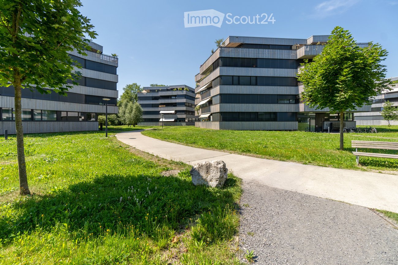 Modern residential complex with contemporary buildings, green lawn, pathways, trees, and a bench.