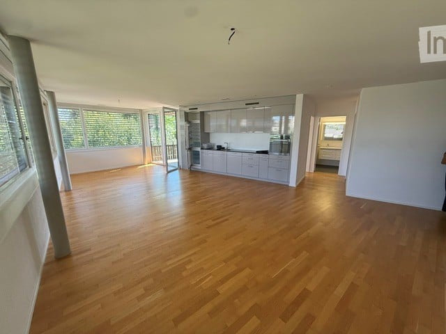 Open-plan living and kitchen area with light wooden flooring, modern kitchen cabinets, and large windows leading to a balcony.