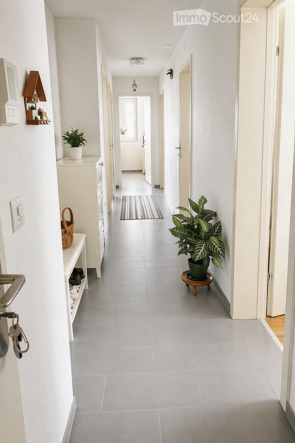 Clean, modern hallway with white walls, tiled floor, and several doors leading to different rooms. Features include a small shelf with shoes, a potted plant, and a decorative rug.