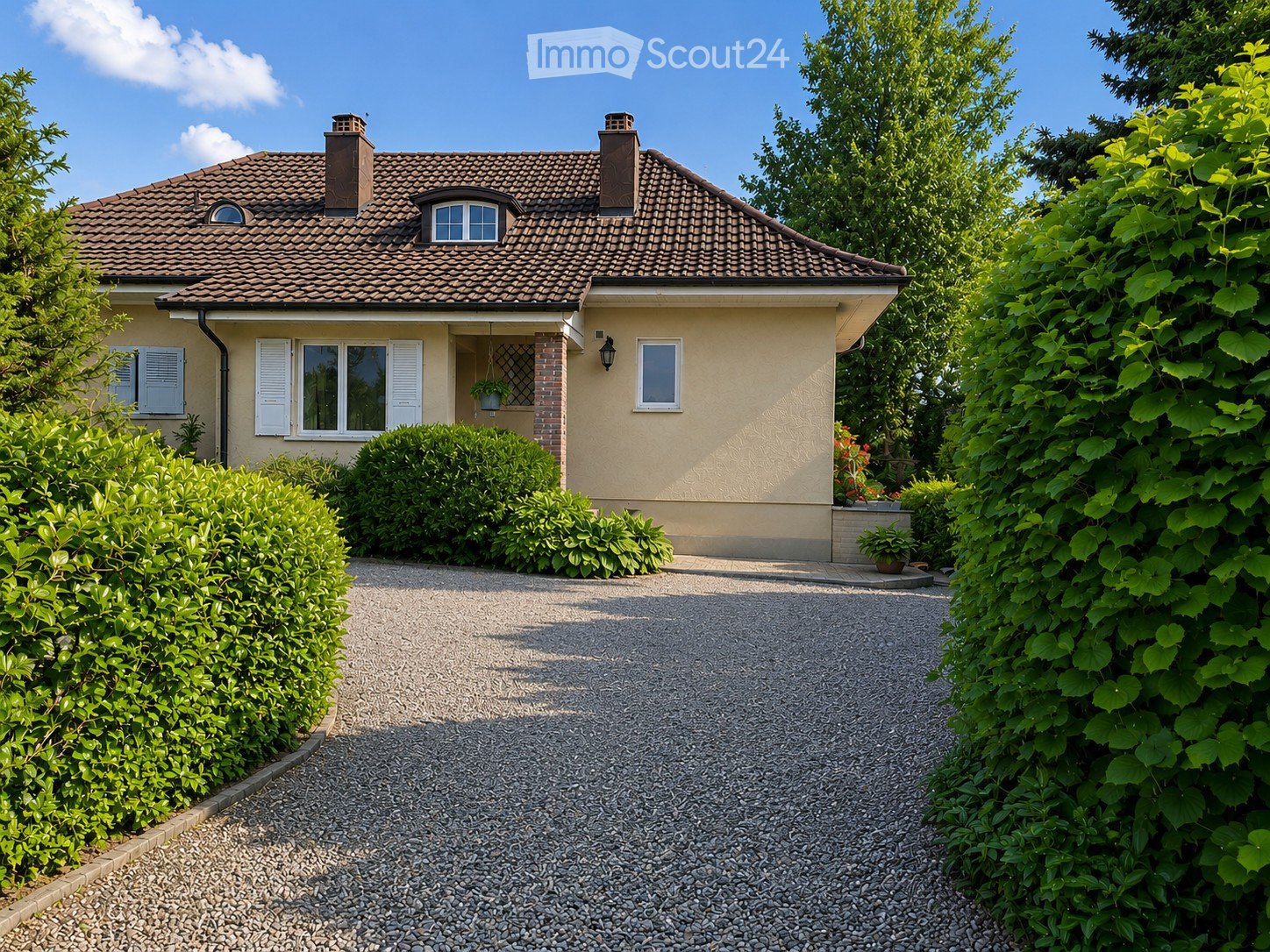 Two-story house with a tiled roof, beige exterior, white shutters, and a gravel driveway surrounded by greenery.