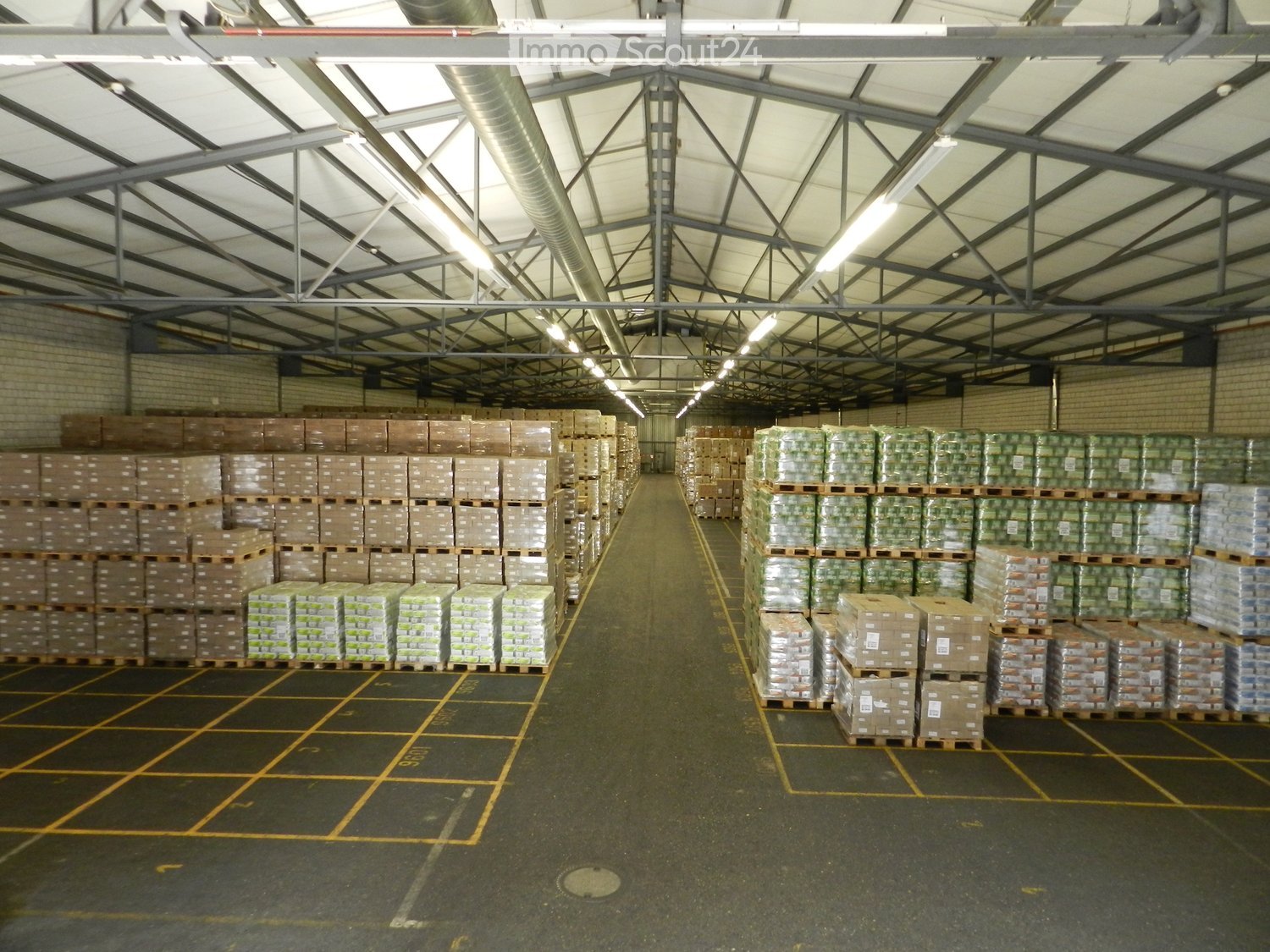 Large industrial warehouse with high ceilings, metal beams, and rows of stacked pallets and boxes on the floor.