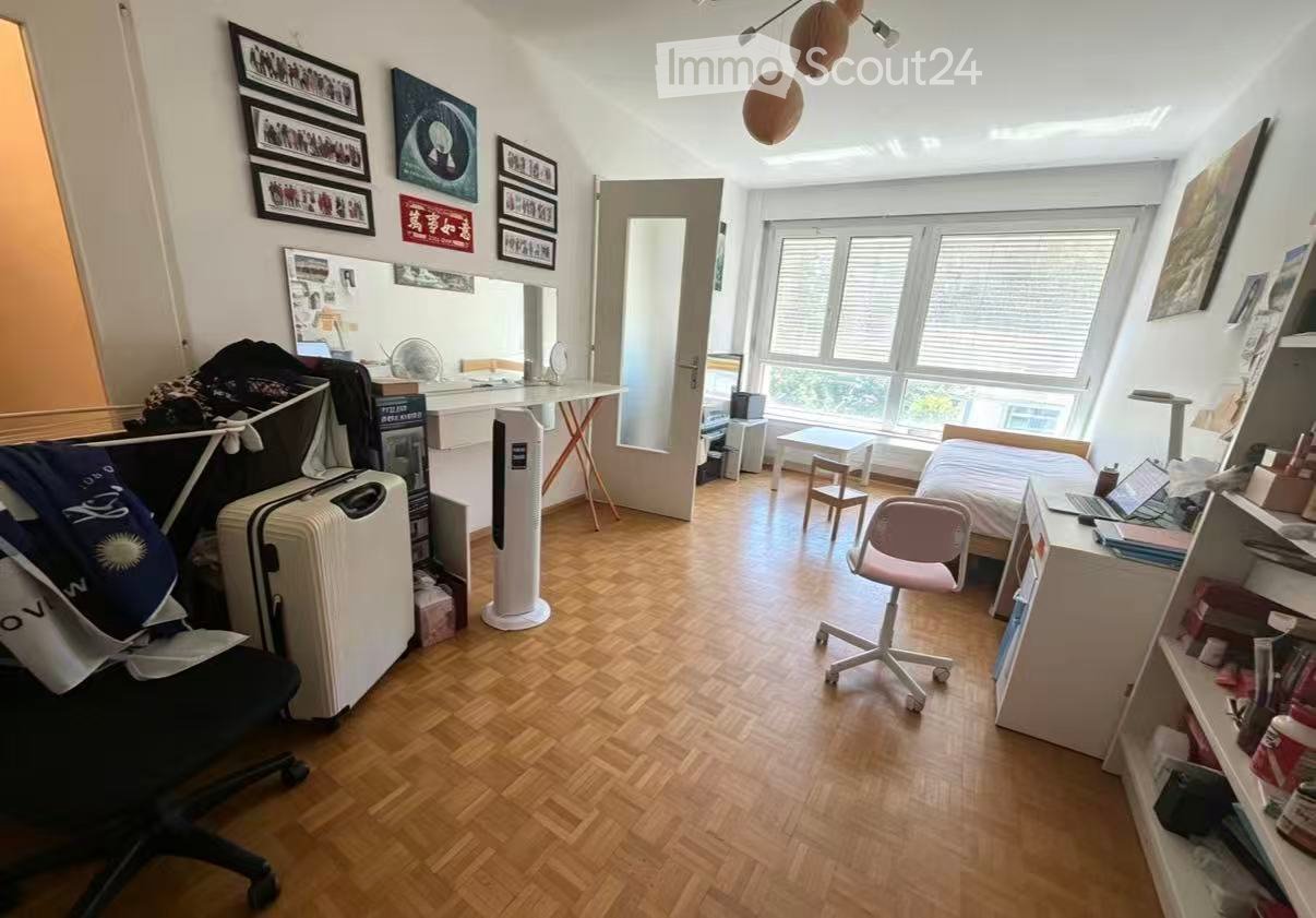 A furnished room with a wooden floor, a bed, a desk with a laptop, a chair, a suitcase, and shelves with various items. The room has large windows with blinds and several picture frames on the walls.