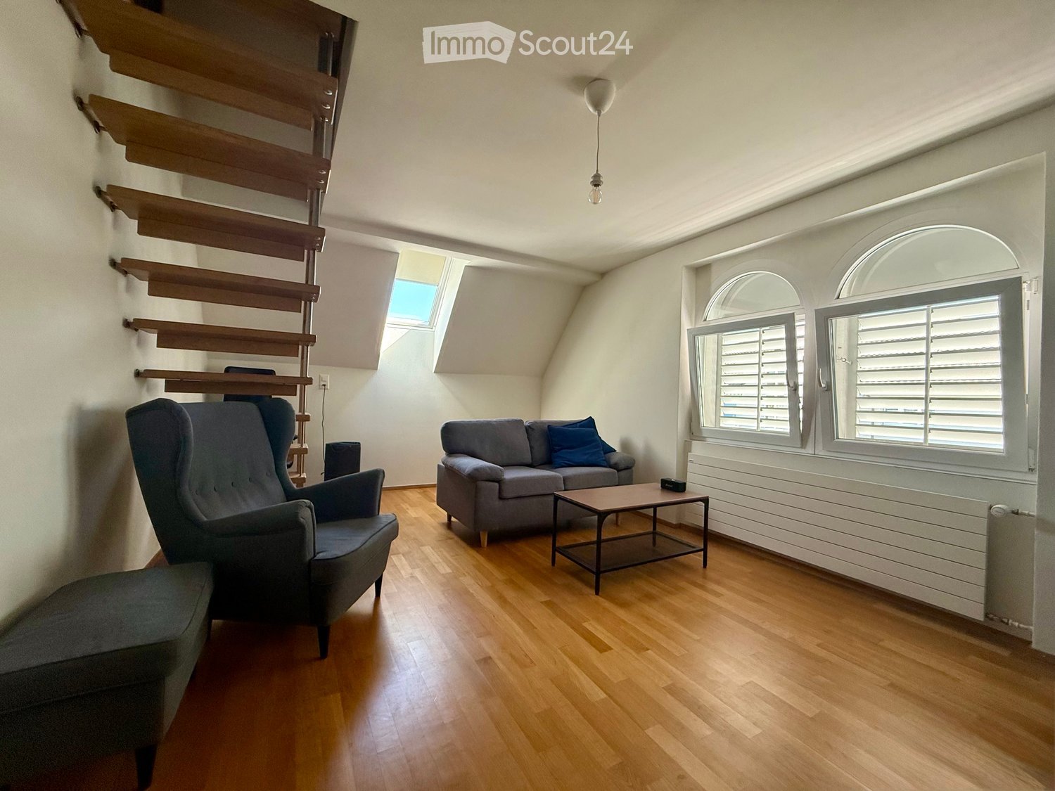 Living room with wooden flooring, grey sofa, armchair, coffee table, and ottoman. Features large windows with shutters and a wooden staircase leading to a loft area.