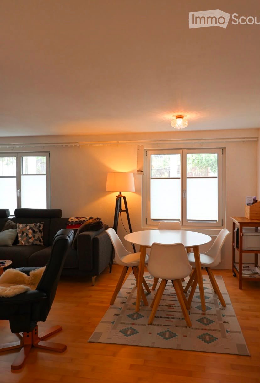 Bright living room with wooden flooring, large windows, a sofa, a round dining table with chairs, a floor lamp, and an ottoman.