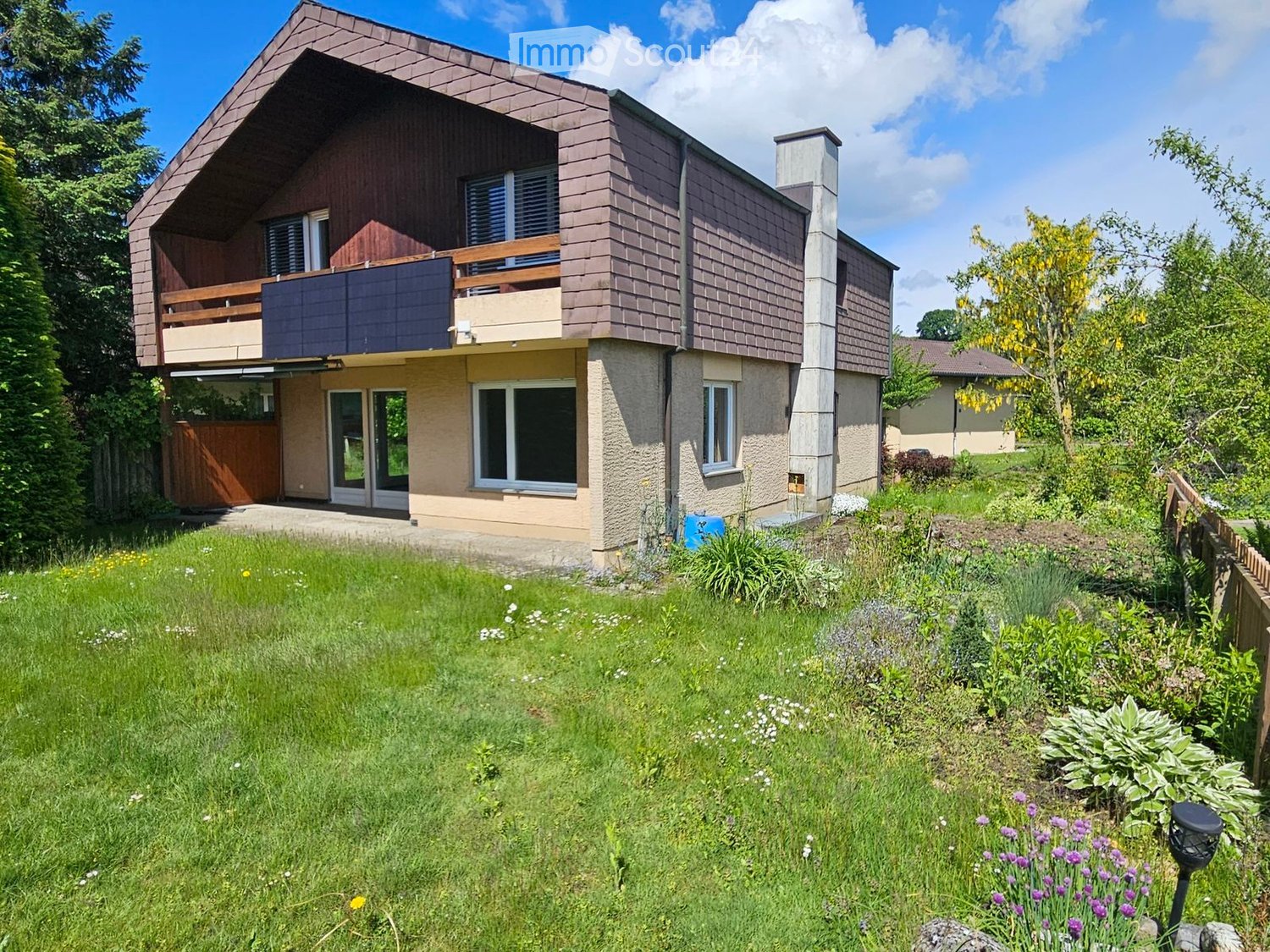 Two-story house with a modern design, featuring a balcony on the upper level, large windows, and a well-maintained garden with various plants and flowers.