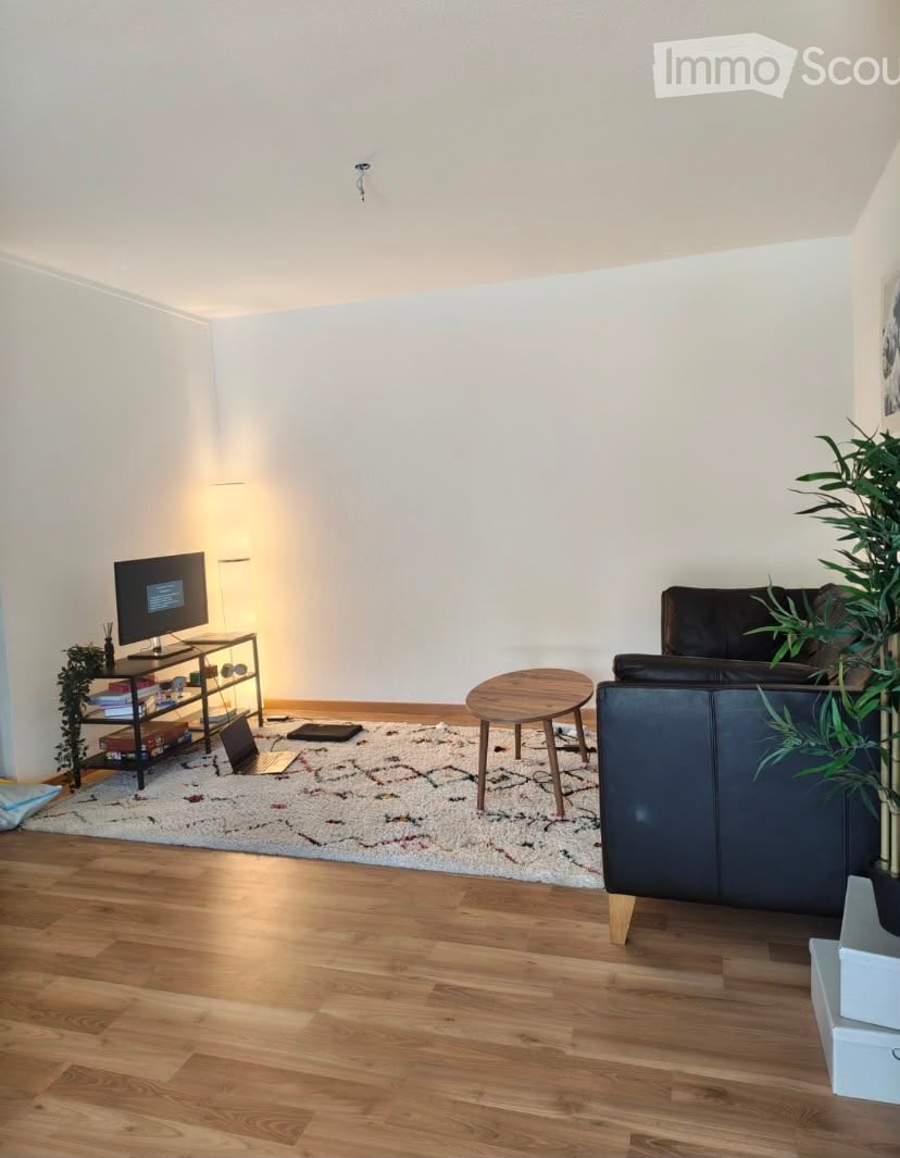 Modern living room with wooden flooring, white walls, a patterned rug, a dark leather sofa, a small wooden coffee table, a TV on a stand, and a floor lamp.