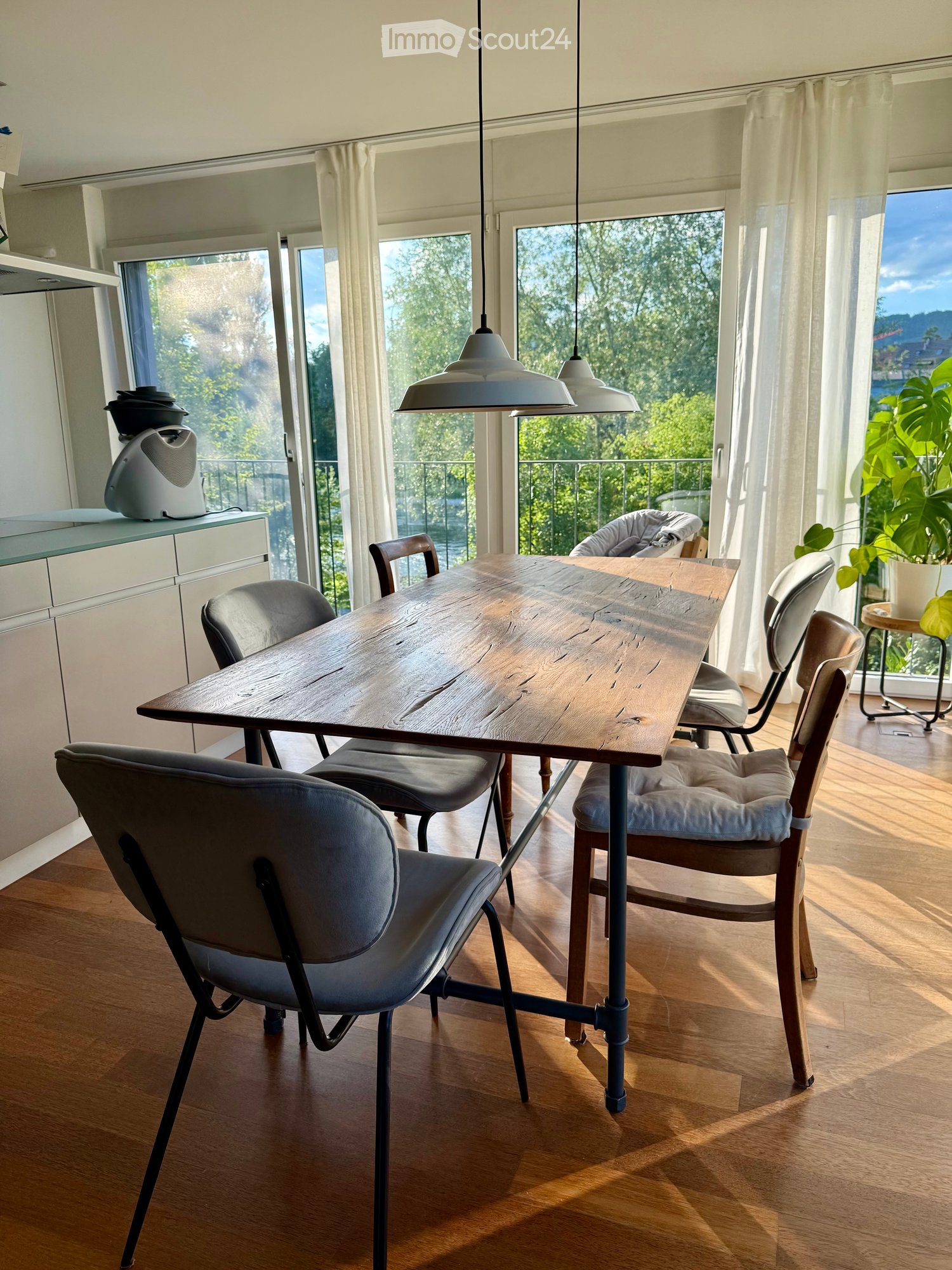 Bright kitchen with wooden table, modern chairs, pendant lamps, and large windows overlooking greenery.
