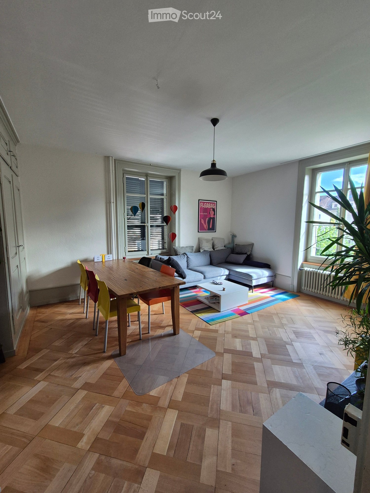 Bright living room with wooden flooring, large windows, a grey sectional sofa, a wooden dining table with colorful chairs, a white coffee table, and a colorful rug.