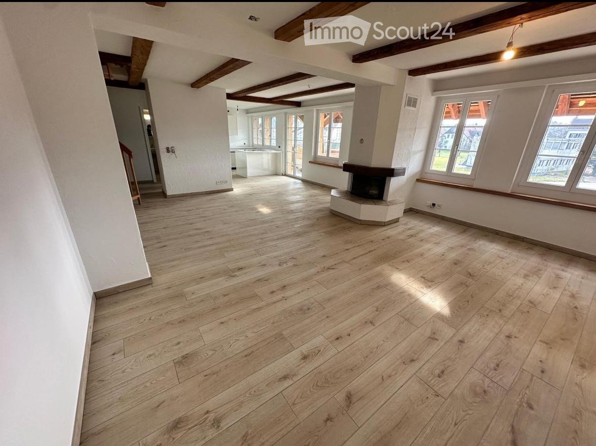 Empty room with wooden flooring, white walls, exposed wooden beams on the ceiling, and a fireplace. Large windows allow natural light to enter.