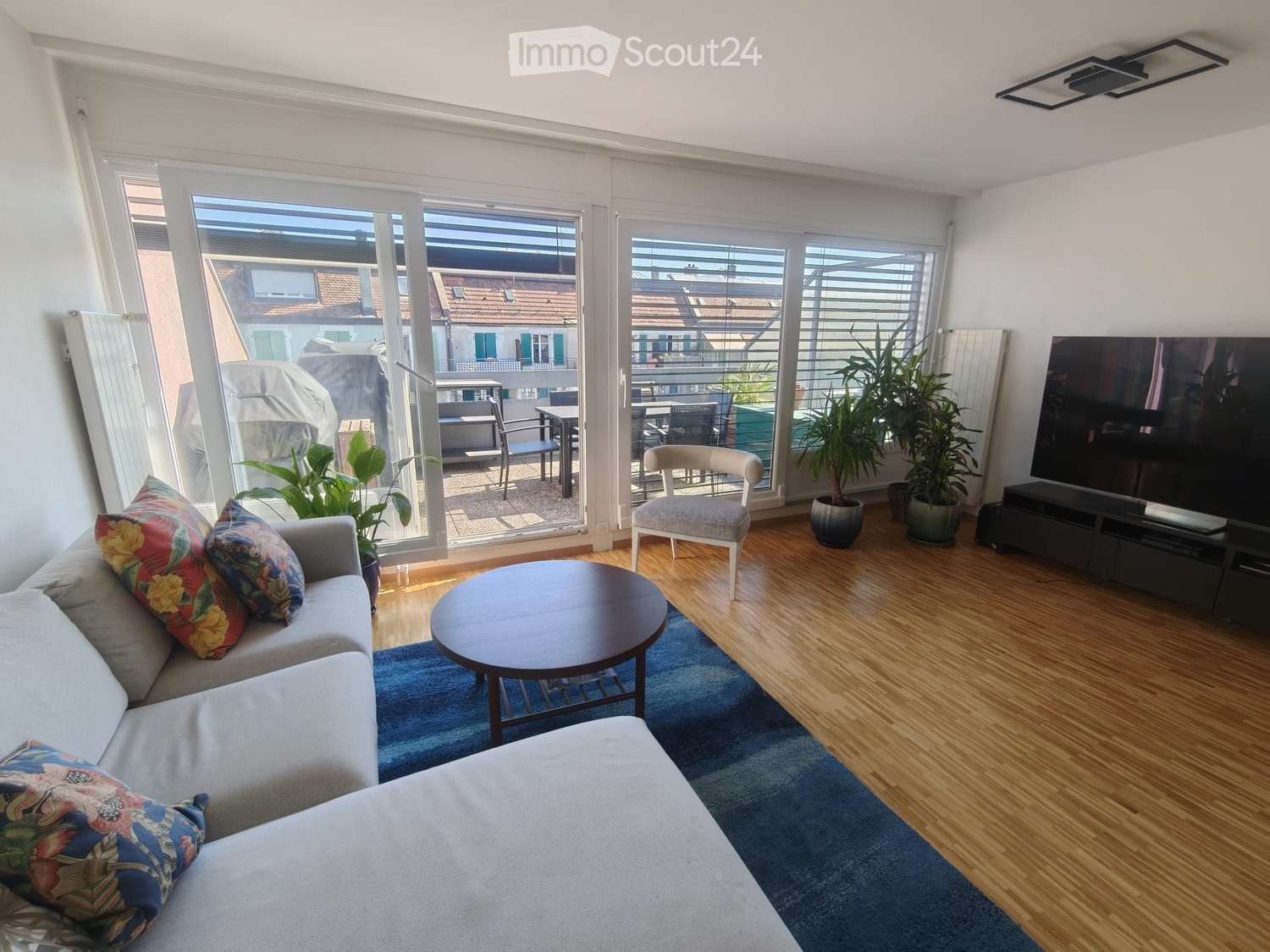 Modern living room with large windows and a balcony, featuring a grey sofa, coffee table, armchair, TV, and potted plants.
