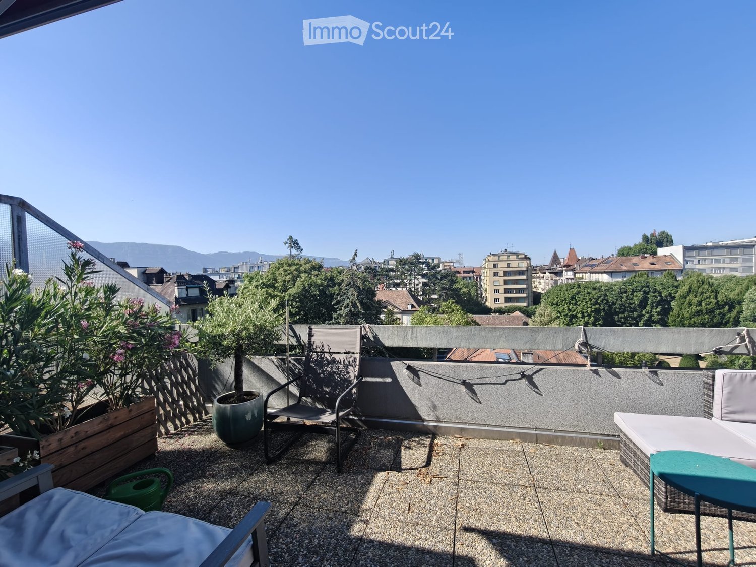 A rooftop terrace with a view of the city and mountains, featuring a sofa, chairs, a small table, and potted plants.