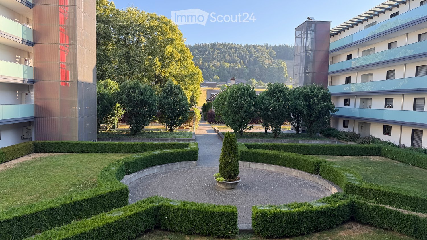 Well-maintained garden area with neatly trimmed hedges, a central tree, and a paved walkway leading to modern apartment buildings.