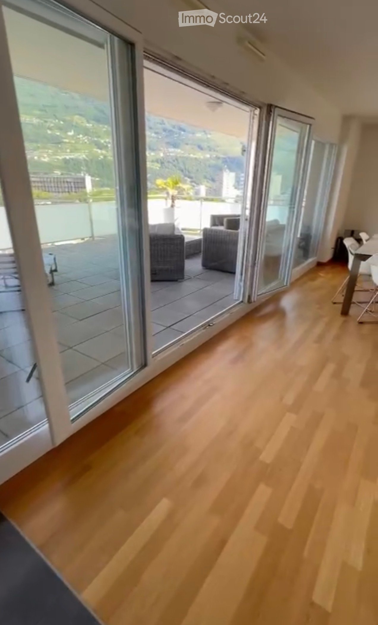 Bright living room with wooden flooring, large glass doors leading to a balcony with outdoor seating and scenic mountain view.