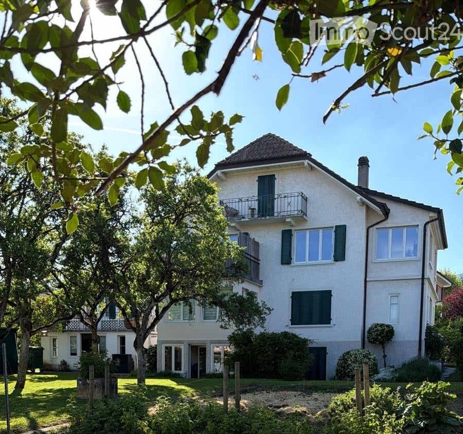 A two-story white house with green shutters, multiple windows, and balconies, surrounded by trees and a well-maintained garden.