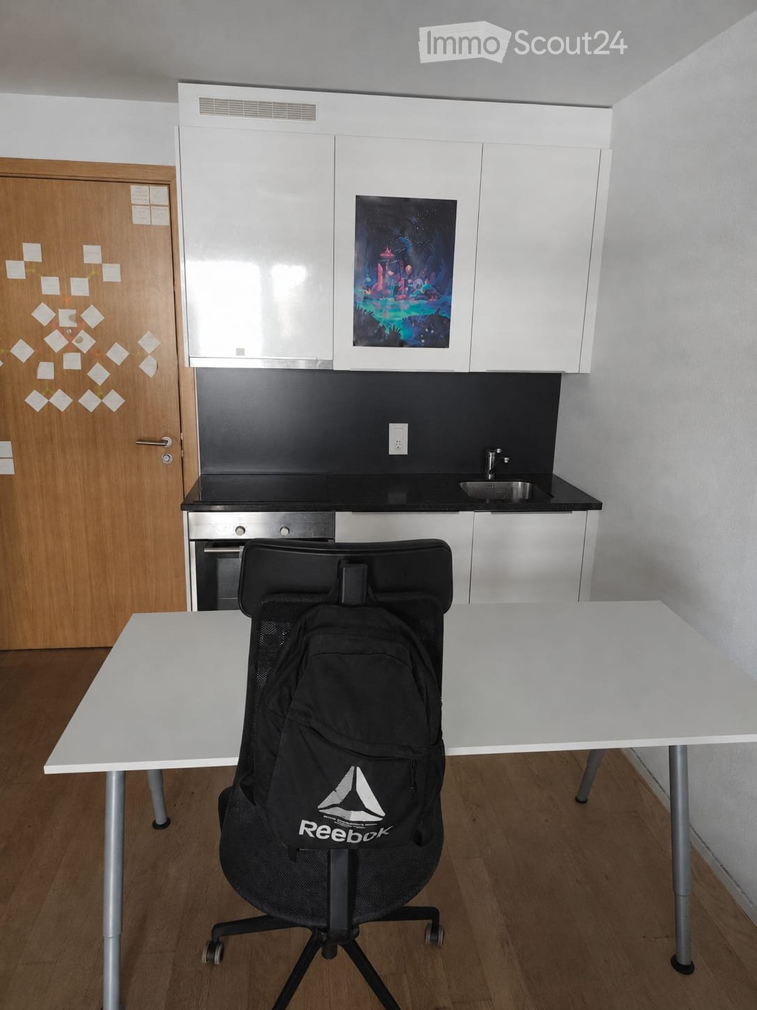 A modern, compact room with a white table, a black Reebok backpack, a black office chair, white cabinets, a sink, and a small oven. The room features a wooden floor and a colorful picture on the wall.