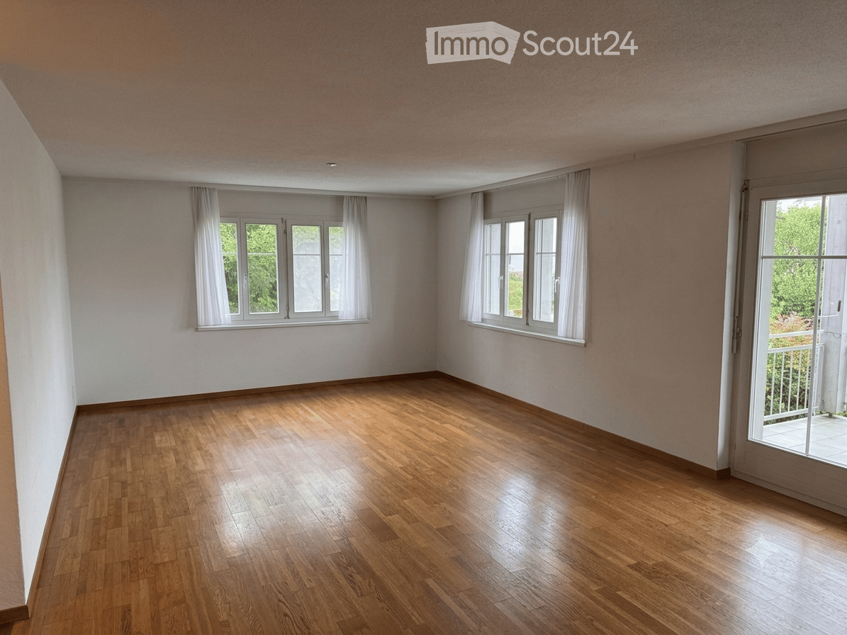 Empty room with wooden flooring, white walls, large windows with white curtains, and a sliding glass door leading to a balcony.