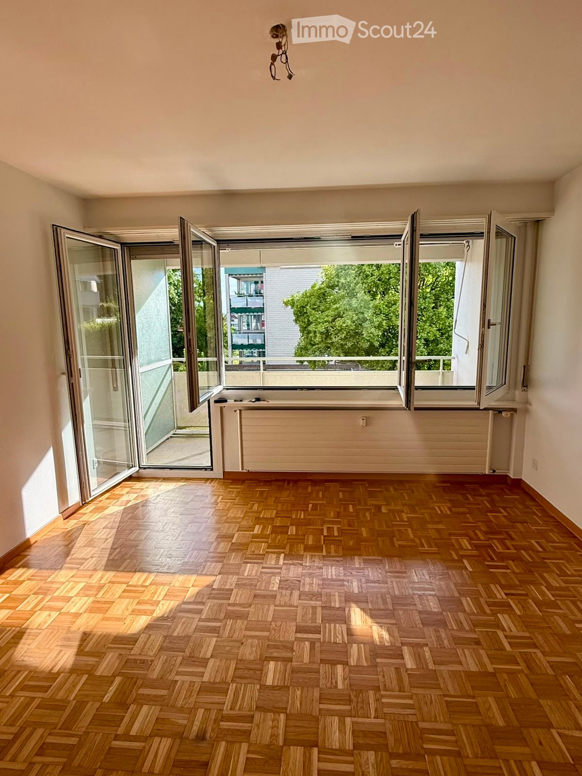 Empty room with wooden herringbone flooring, large windows with sliding glass doors, radiator, and visible electrical outlet.