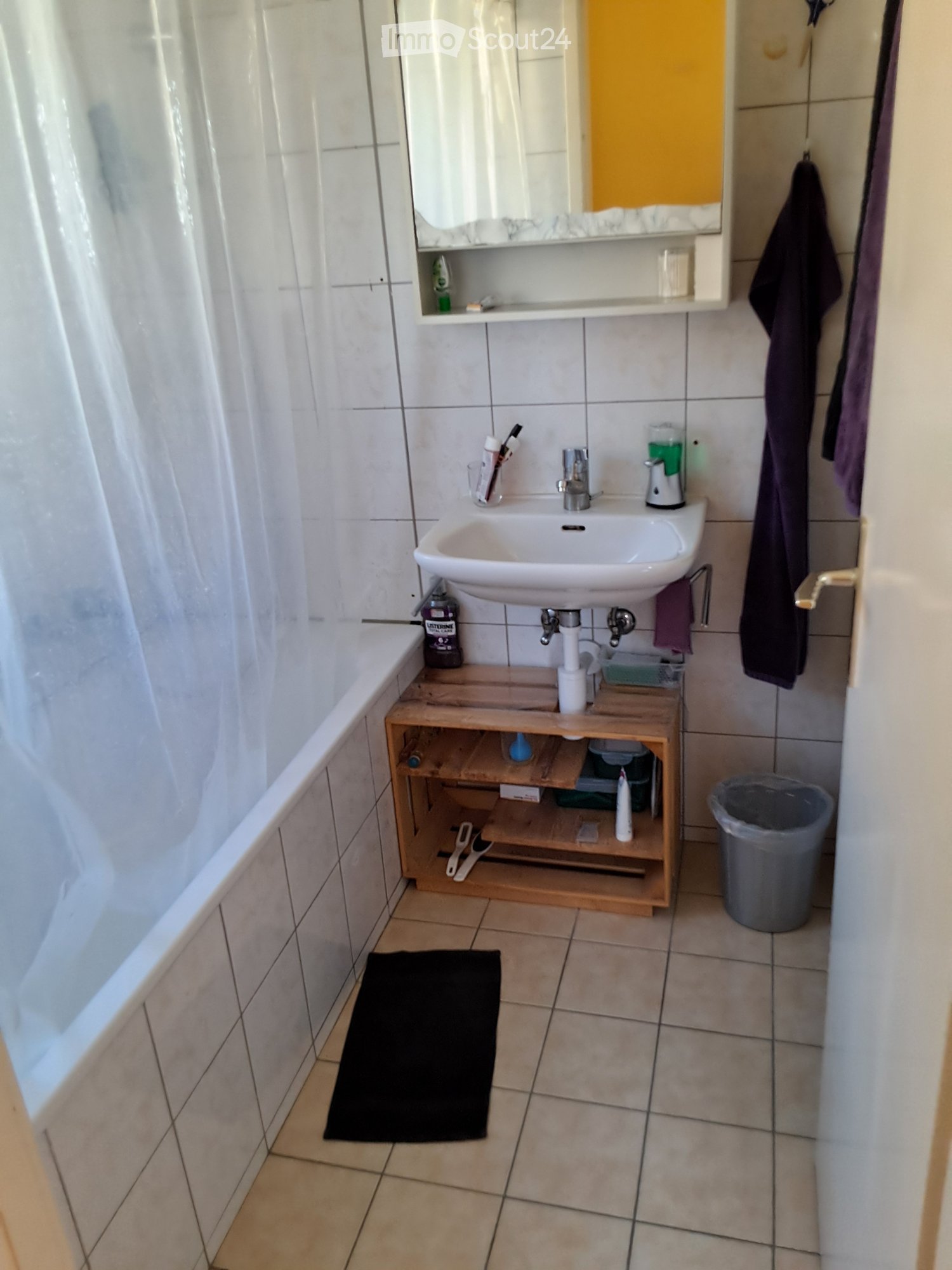 The bathroom features a white sink with a mirror above it, a wooden shelf holding various toiletries, a black towel on the floor, and a trash bin in the corner.