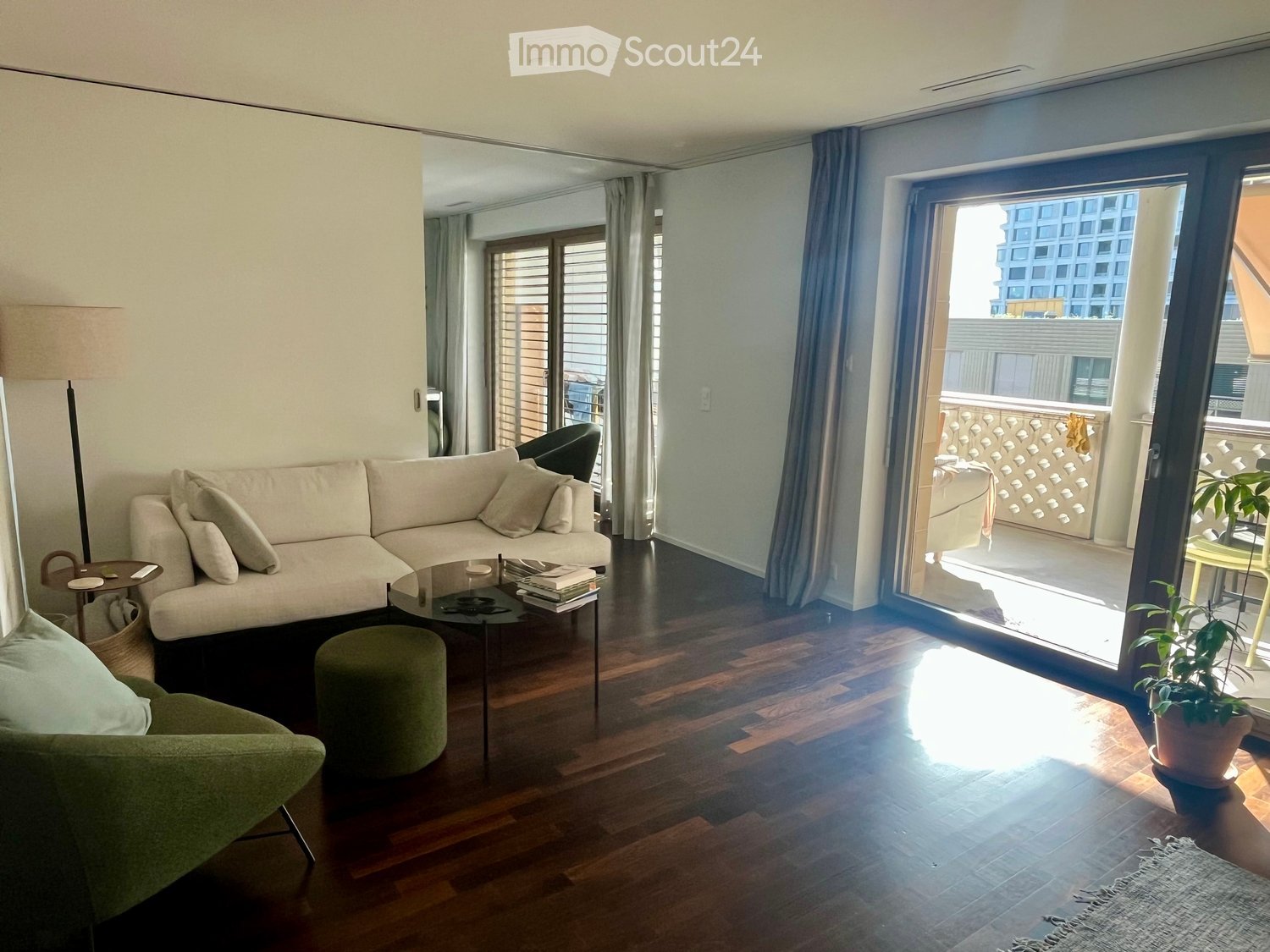 Modern living room with a light-colored sectional sofa, glass coffee table, green stool, and a balcony with outdoor seating visible through large glass doors.