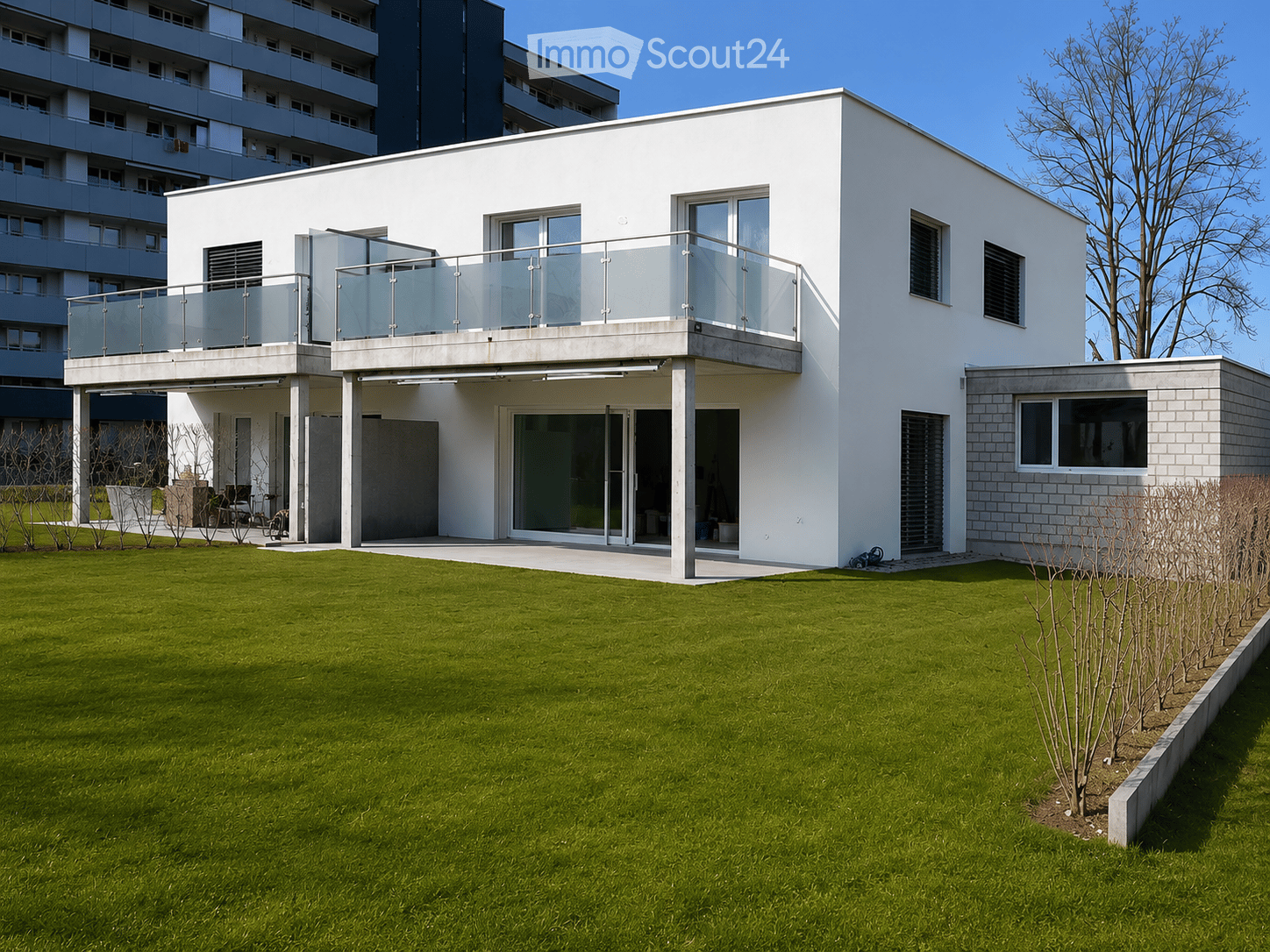 Modern two-story house with white exterior, large windows, and a balcony with glass railing. The house features a well-maintained lawn and is situated in a residential area with neighboring buildings.