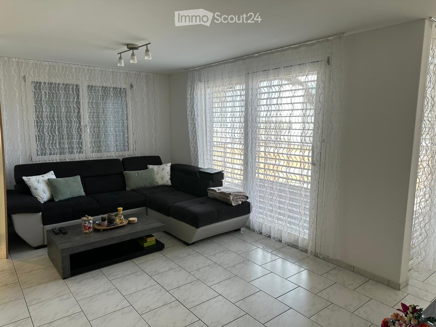 Modern living room with a black sectional sofa, grey coffee table, decorative items, and large windows with sheer curtains.