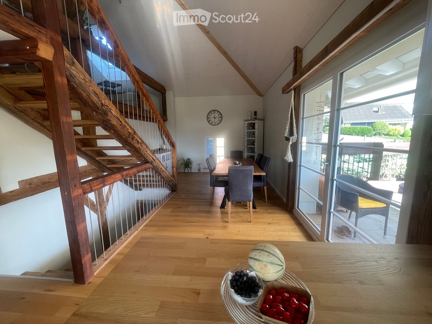 Bright, open-plan living and dining area with wooden flooring, a staircase leading upstairs, and large windows offering a view of the garden. The dining area features a wooden table with chairs, and a fruit basket is placed on the coffee table.