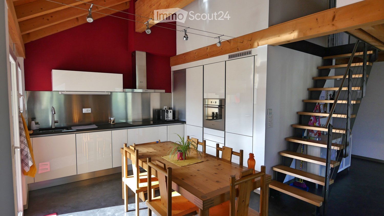 Modern kitchen with white cabinets, stainless steel appliances, a wooden dining table with chairs, and a staircase leading to another level.