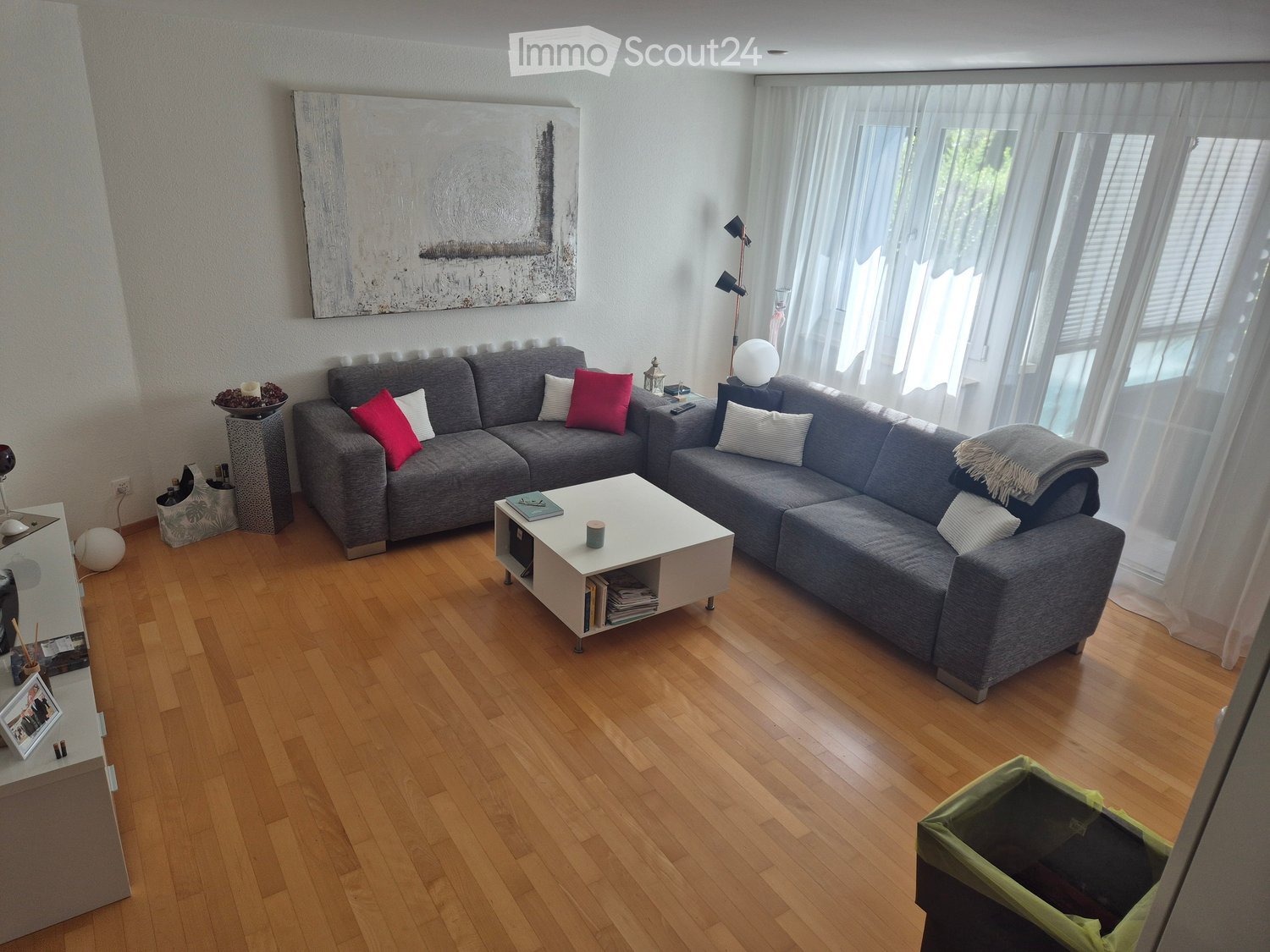 Modern living room with wooden flooring, large grey sectional sofa, white coffee table, floor lamp, and large windows with sheer curtains.