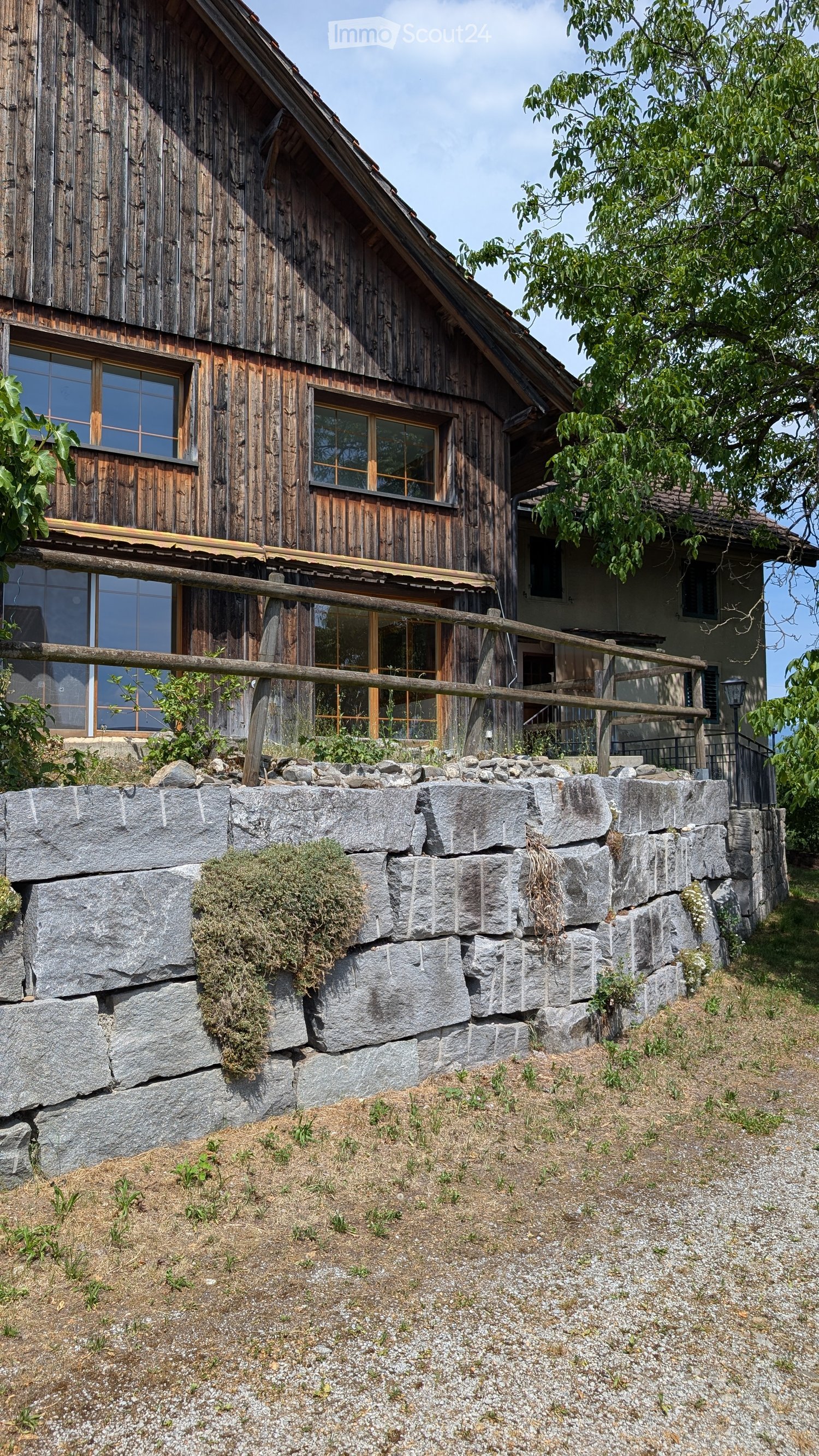 A rustic wooden house with a stone retaining wall in front, featuring large windows and a wooden balcony.