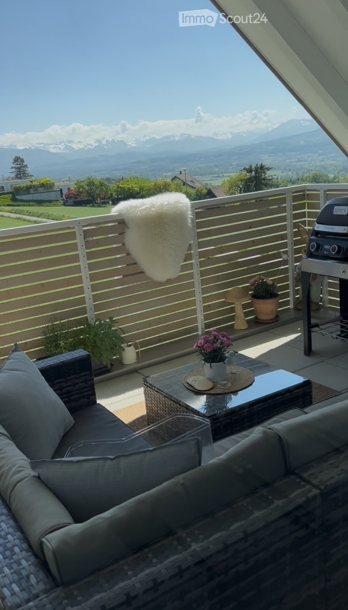 Balcony with a view of mountains, featuring a wicker sofa, a small table with flower pots, and a grill.