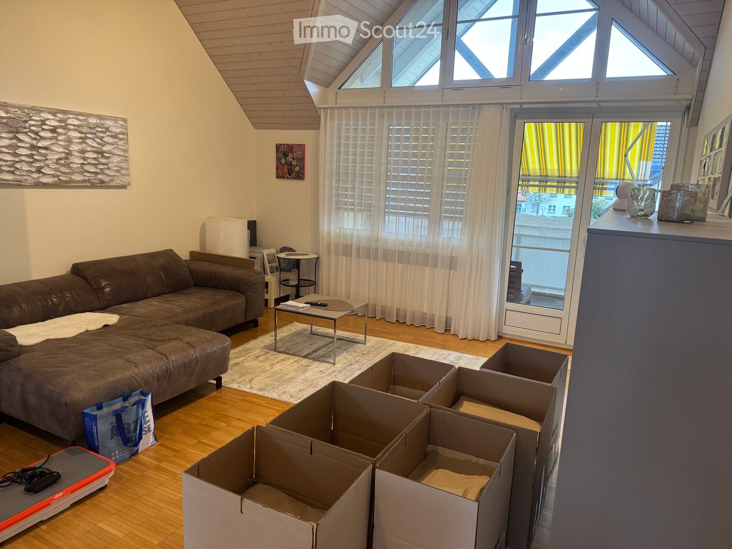 Living room with a brown leather sofa, coffee table, side table, and large windows with curtains. Several cardboard boxes are scattered on the floor.