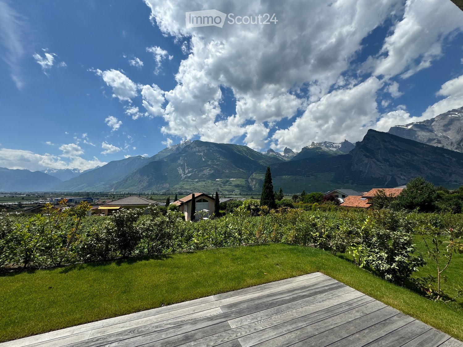 A well-maintained garden with a wooden deck, green grass, and surrounding bushes, overlooking a scenic mountain range under a partly cloudy sky.