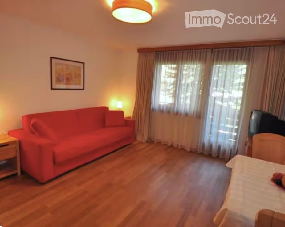 A cozy living room with a red sofa, wooden floor, a small table with a lamp, a television, and a picture frame on the wall. Large windows with curtains let in natural light.
