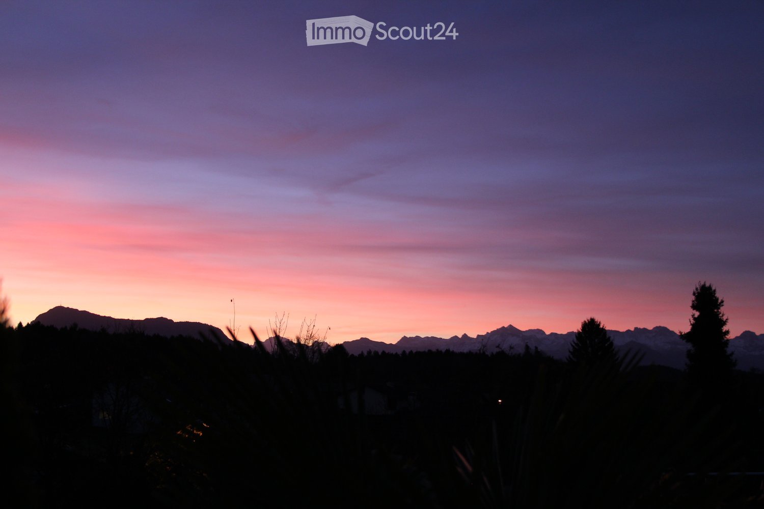 A serene outdoor view at sunset, featuring a mountain range silhouetted against a colorful sky with hues of pink, purple, and orange. The foreground includes dark silhouettes of trees and shrubs.
