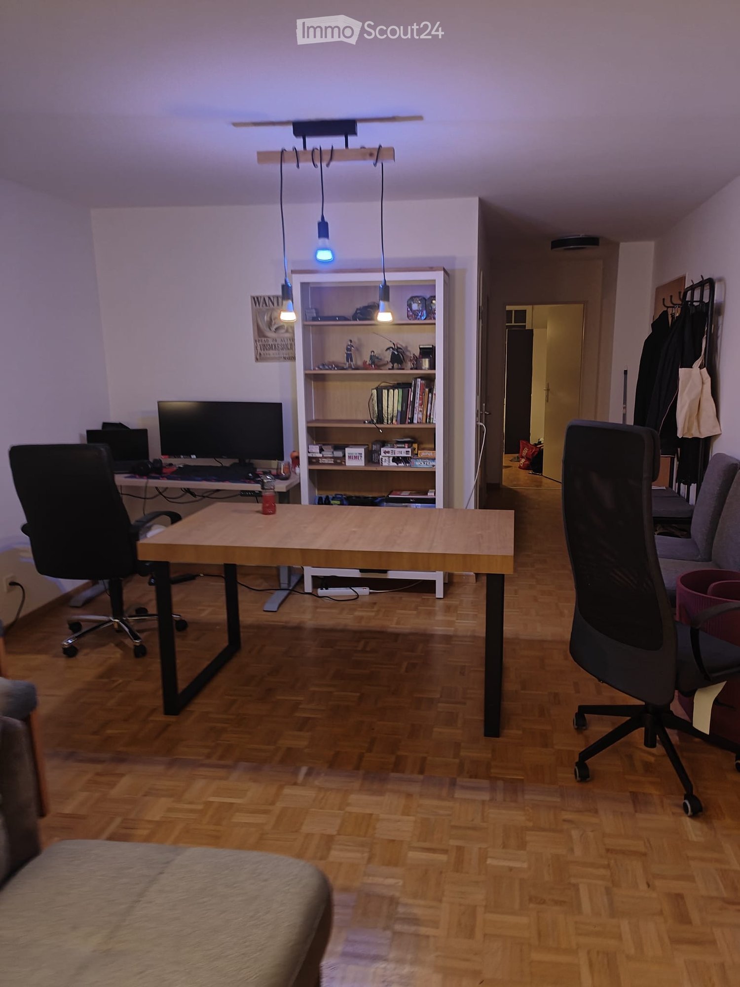 A modern, well-lit room with wooden flooring, a large wooden table, two office chairs, a sofa, a shelf with books and decorative items, and a computer setup with a screen and keyboard.