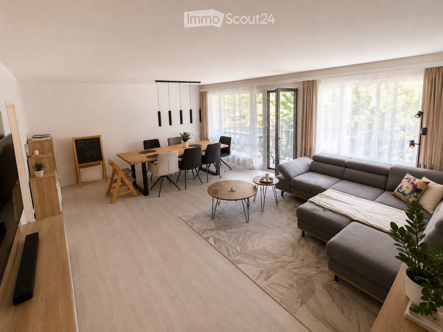 Modern living and dining area with a grey sectional sofa, wooden coffee table, dining table with chairs, and pendant lighting.