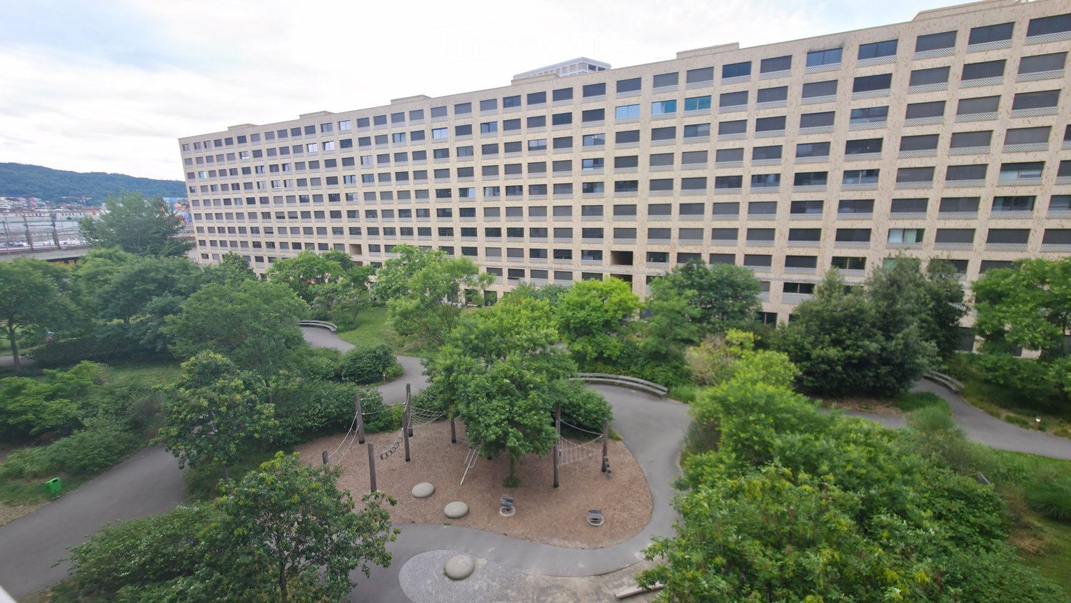 Aerial view of a large residential building surrounded by a landscaped garden area with pathways, trees, and a small playground area with a volleyball net and stone elements.