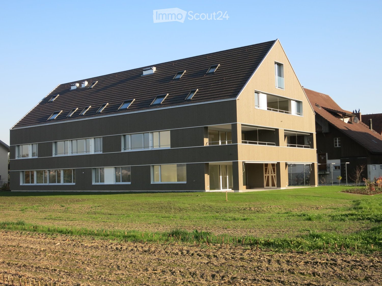 Modern two-story building with a sloped roof, large windows, and balconies, surrounded by a grassy lawn.