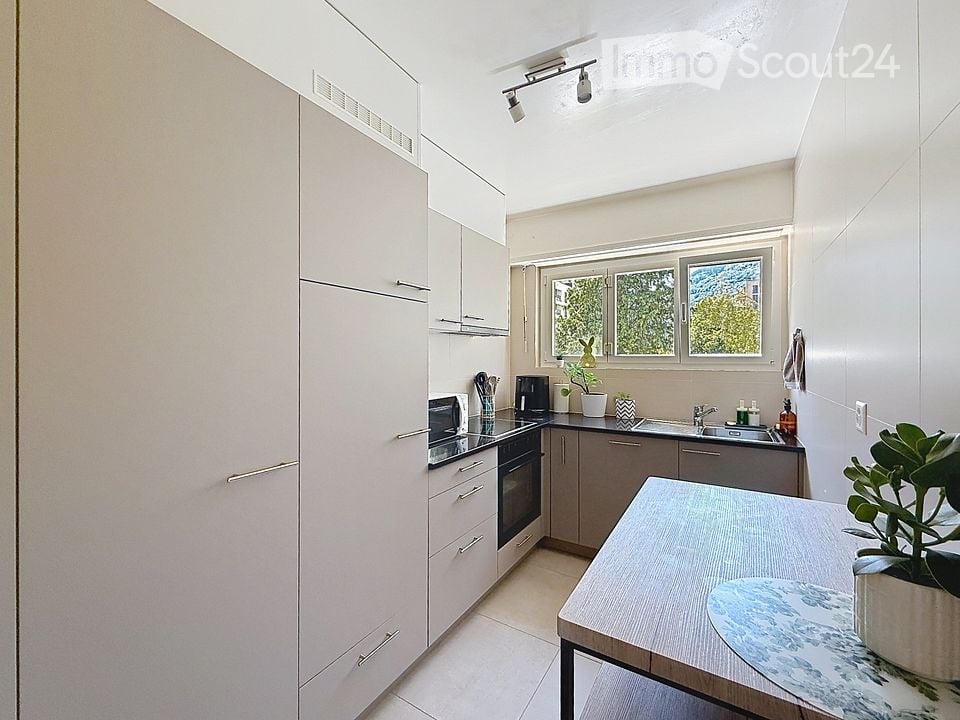 Modern kitchen with white cabinets, stainless steel appliances, and a central island table with a potted plant. The space features a window with a view of greenery outside.