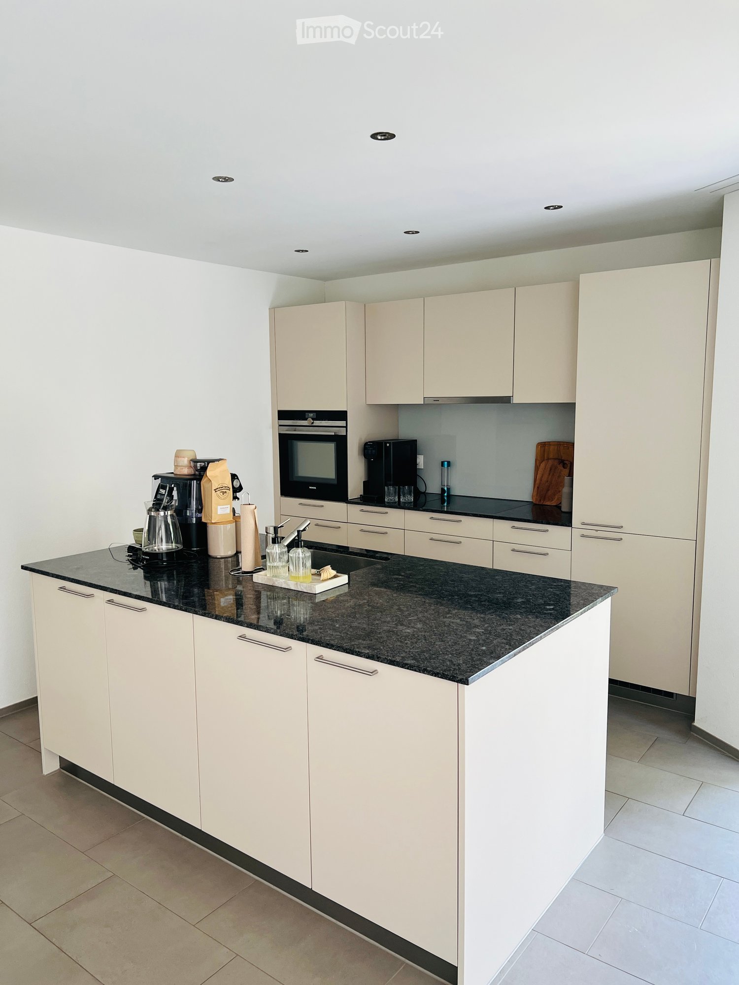 Modern kitchen with light-colored cabinets, granite countertop, and various appliances including a coffee maker, oven, and cutting board.