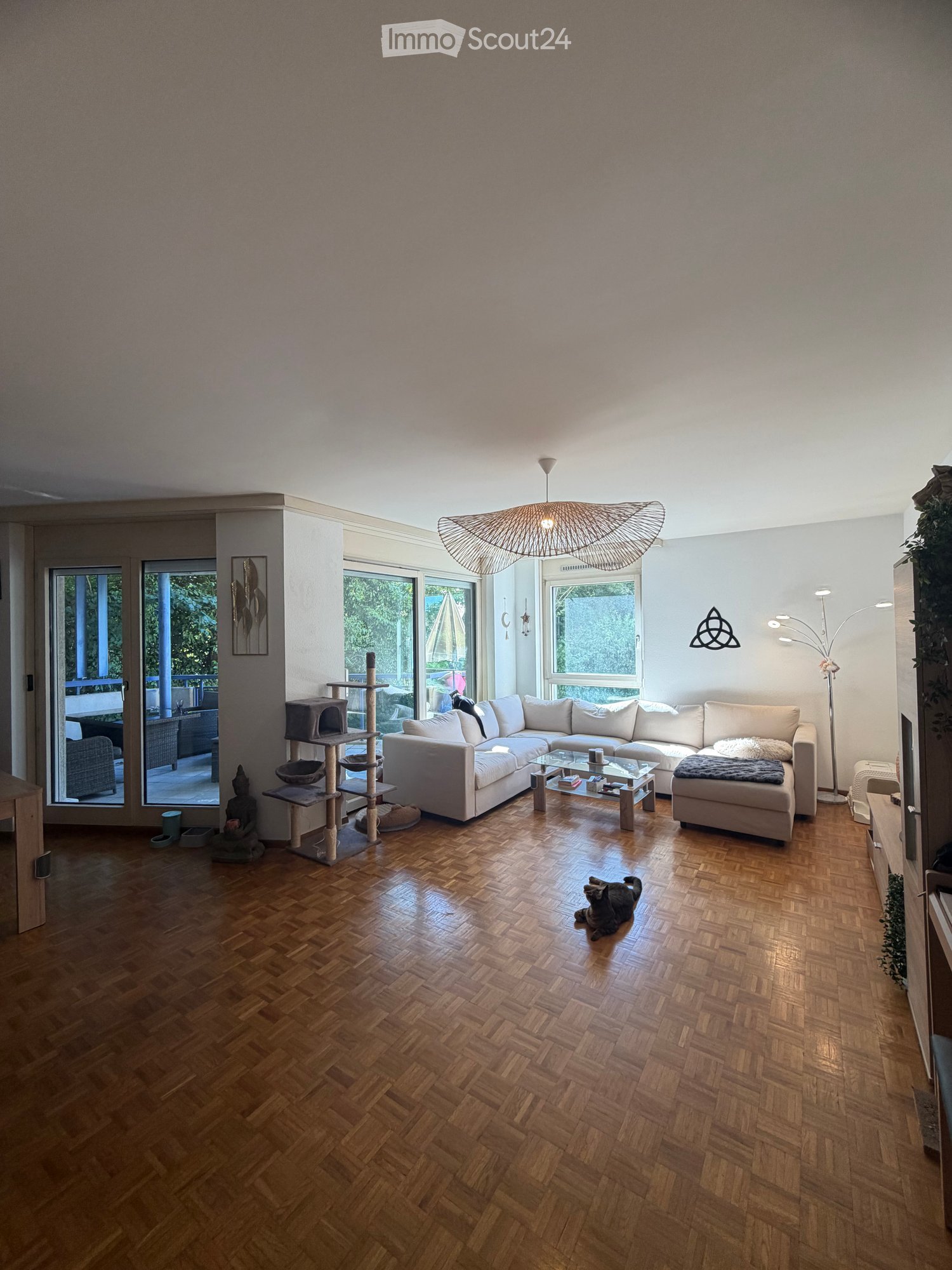 Bright living room with wooden herringbone flooring, large windows, and a modern hanging light fixture. Features a white sectional sofa, glass coffee table, cat tree, and decorative items.