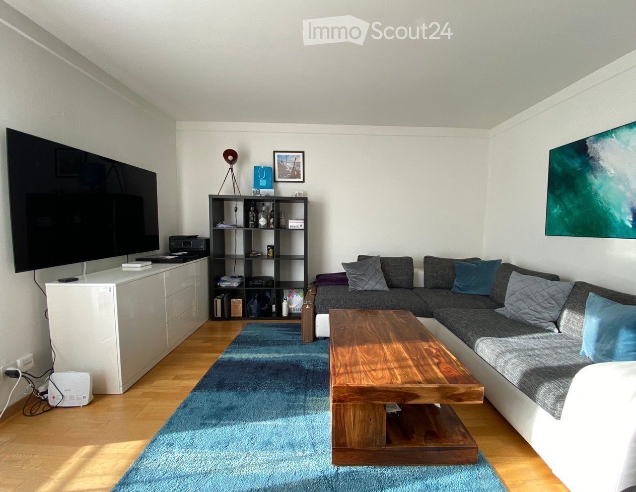 Modern living room with a grey sectional sofa, wooden coffee table, blue rug, TV, shelf with various items, and artwork on the walls.