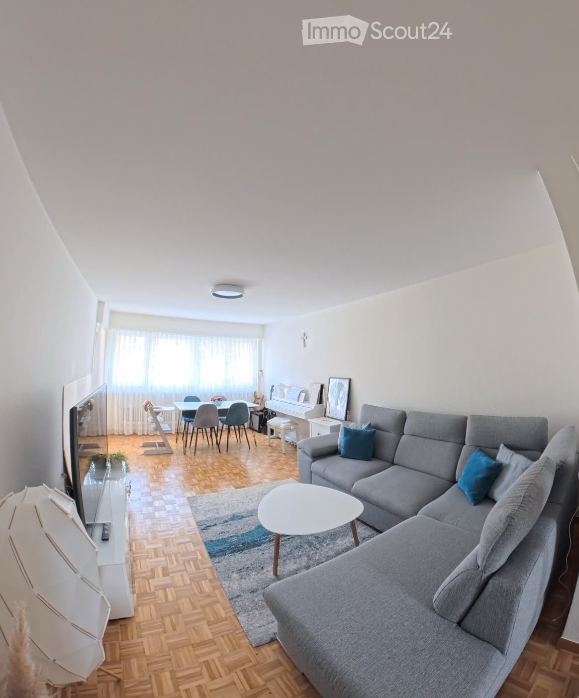 Modern living room with grey sectional sofa, coffee table, TV, dining area with table and chairs, piano, and a small plant. The room features wooden flooring and large windows with curtains.