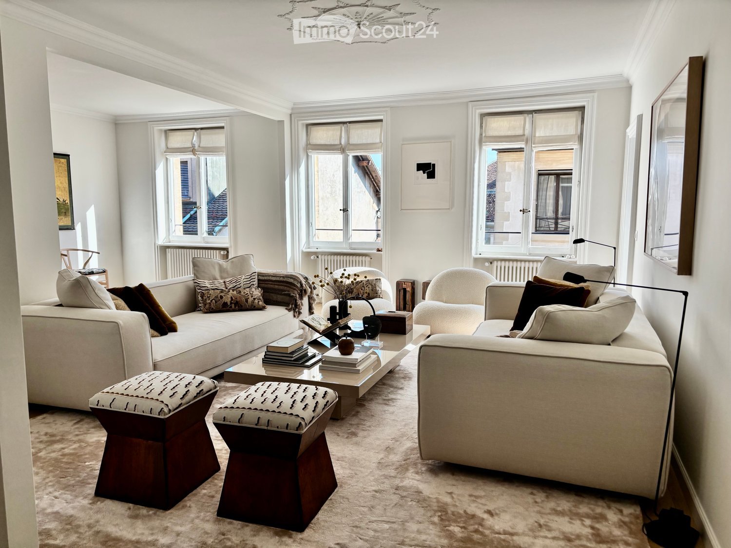 Elegant living room with white walls, large windows, cream-colored sofa, coffee table, two wooden stools, and a small side table with a vase.